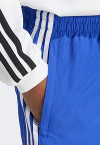 Osoba v modrých sportovních šortkách s bílými a modrými pruhy, ruka ve kapsě, a v bílé dlouhé rukávě s černými pruhy na rukávu.