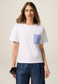 Maglietta bianca in cotone con taschino a righe blu e bianche, caratterizzata da dettagli frangiati. Abbinata a pantaloni in denim blu.