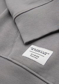 Manche de sweat-shirt en tissu gris avec une étiquette blanche cousue portant l'inscription "VOTRE SEULE LIMITE EST VOTRE ESPRIT" et "OMBRE depuis 1985"