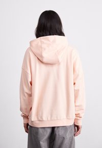 Ljusrosa hoodie med mjuk textur, oversized design och huva. Har sänkta axlar och ribbad nederkant. Bakvy visas.