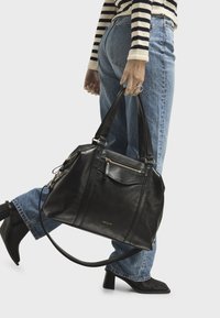 Borsa a tracolla in pelle nera con accessori dorati, tasche con zip, doppi manici e finitura testurizzata, tenuta da una persona con jeans blu.