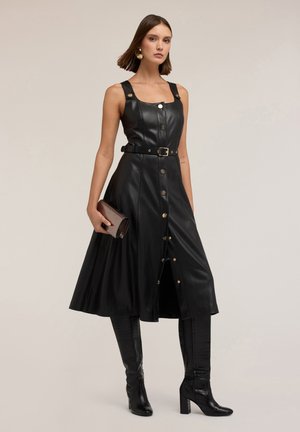 Schwarzes Lederkleid mit eng anliegendem Oberteil, breiten Trägern, goldenen Knöpfen, einem taillierten Gürtel und einem schwingenden Rock; kombiniert mit kniehohen Stiefeln.