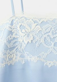 Tissu bleu clair avec superposition de dentelle florale blanche et fines bretelles bleues, montrant la texture et le motif détaillés de la dentelle.