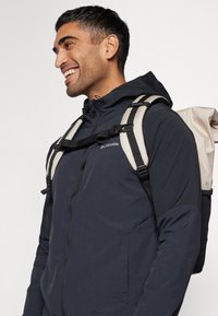 Men's navy jacket with a zippered front and hood, featuring contrasting beige backpack straps and a textured fabric.