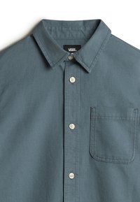 Camicia button-up in tessuto verde scuro, con colletto classico, tre bottoni e una tasca sul petto sinistro con dettagli cuciti.