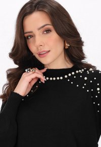 Maglione nero con applicazioni di perle sulle spalle. La modella ha i capelli ondulati e indossa orecchini a cerchio d'oro e anelli d'argento.