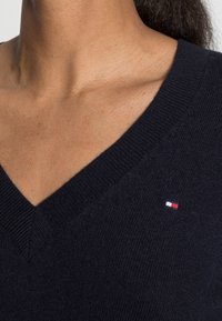 Marinblå v-ringad tröja i mjukt stickat material, med en liten röd, vit och blå logotyp på vänster sida nära halsringningen.