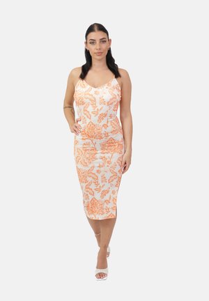 Femme aux cheveux foncés portant une robe midi blanche avec des motifs floraux orange et des sandales blanches à talons, debout avec une main sur la hanche.