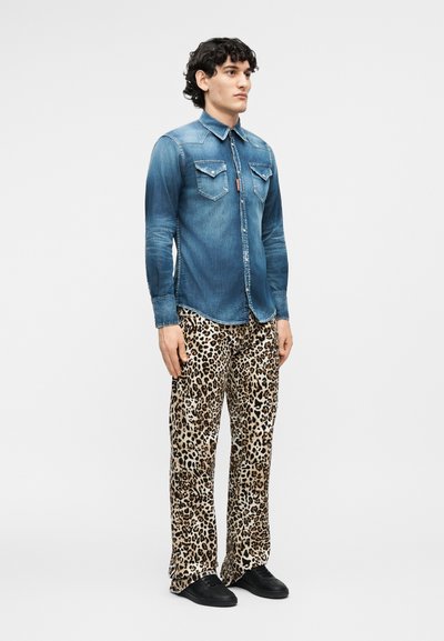 Νεαρός άνδρας στέκεται φορώντας ξεθωριασμένο μπλε πουκάμισο από τζιν με τσέπες στο στήθος και φαρδιά παντελόνια με λεοπάρ σχέδιο, συνδυασμένα με μαύρα παπούτσια.