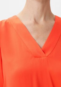 Blouse à col en V de couleur orange vif, fabriquée en tissu lisse. Présente un léger éclat, avec une finition soignée au niveau de l'encolure et des épaules.