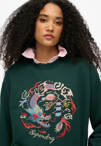 Donkergroene sweatshirt met levendige geborduurde ontwerpen van een draak, wolken en een pagode, met een ronde halslijn en een zachte stoftextuur.