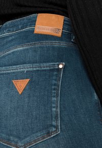 Vaqueros de mezclilla azul con un parche de cuero marrón que dice "GUESS ICON" y un logo de triángulo en el bolsillo trasero. Detalles de costura visibles.