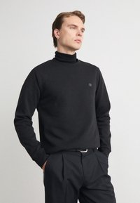 Black turtleneck sweater in a soft fabric, featuring long sleeves and a subtle logo on the chest. Paired with dark trousers.