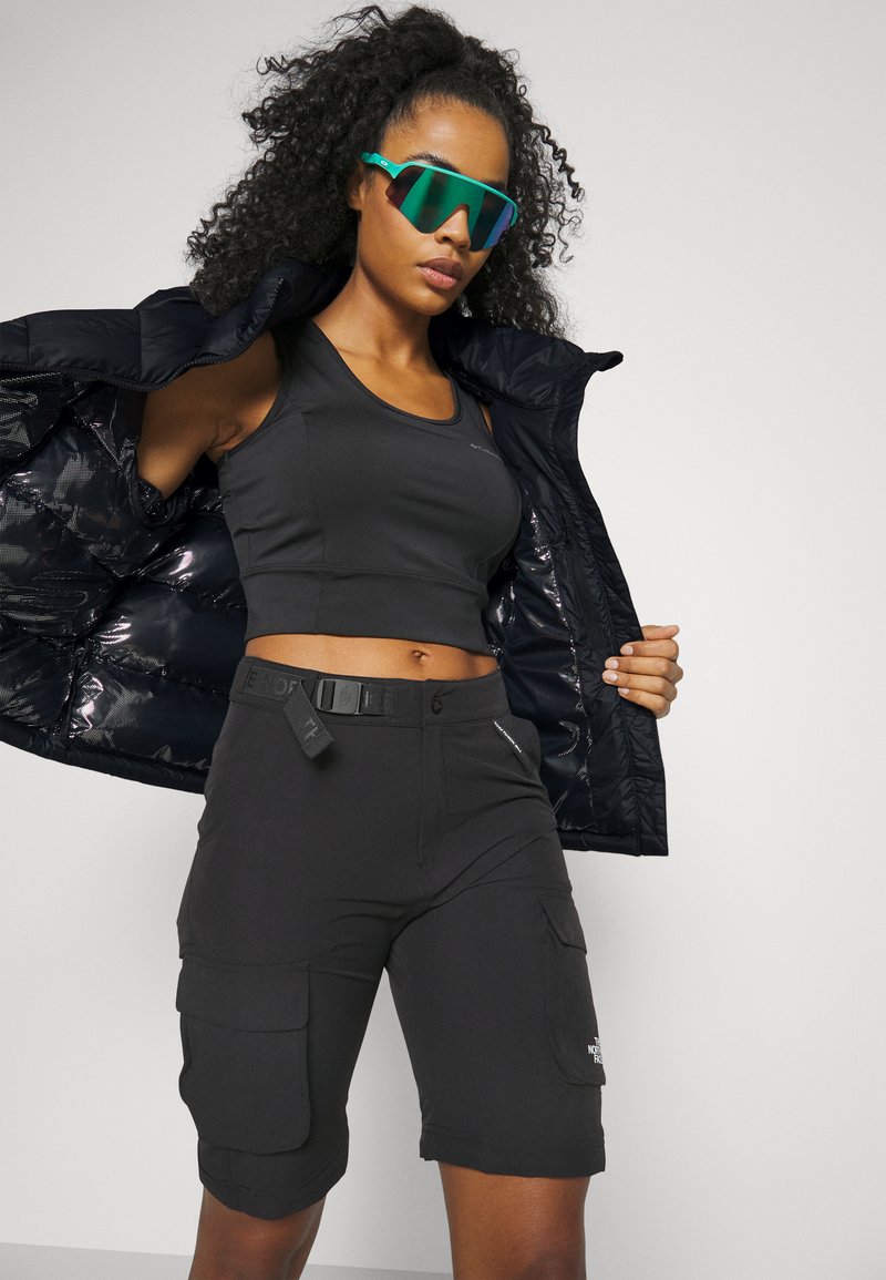 Mujer vestida con un top corto negro, pantalones cortos cargo negros con cinturón, chaqueta negra abierta y gafas de sol deportivas reflectantes verdes, contra un fondo liso.