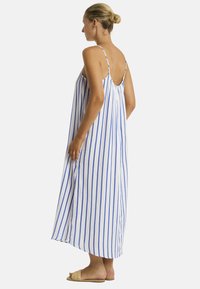 Una donna sta in piedi di lato, indossando un vestito maxi senza maniche a righe blu e bianche, abbinato a sandali chiari.