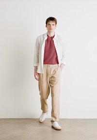Lett, hvit skjorte over en maroon teksturert skjorte, kombinert med beige bukser med snøring og hvite sneakers, mot en nøytral bakgrunn.