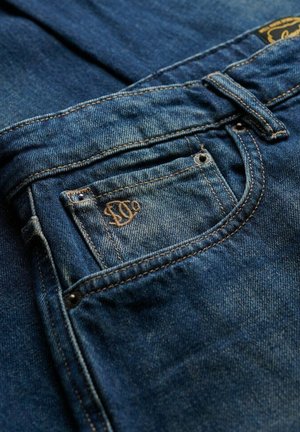 Granatowe dżinsy z ciemnego denimu z tylną kieszenią z embosowanym logo i kontrastującymi szwami. Tkanina ma nieco wyblakłą fakturę.