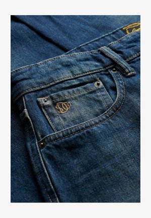 Granatowe dżinsy z ciemnego denimu z tylną kieszenią z embosowanym logo i kontrastującymi szwami. Tkanina ma nieco wyblakłą fakturę.