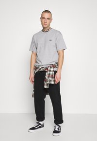 Camiseta de algodón gris con un pequeño logo, combinada con una camisa de franela de cuadros atada a la cintura, pantalones negros y zapatillas negras con detalles en blanco.