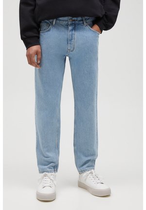 Osoba nosząca jasnoniebieskie jeansy o prostych nogawkach, białe trampki i czarną bluzkę z długim rękawem, z jedną ręką w kieszeni.