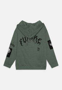 Sweat à capuche vert avec cordon de serrage, arborant des lettres graphiques noires, des patchs texturés sur les manches et une coupe décontractée.
