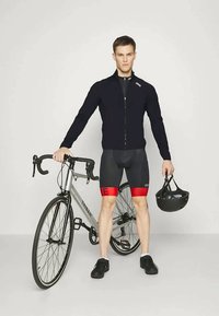 Ciclista in una giacca nera con zip e logo bianco, pantaloni corto con dettagli rossi e scarpe nere, che tiene un casco nero accanto a una bicicletta da strada grigia.