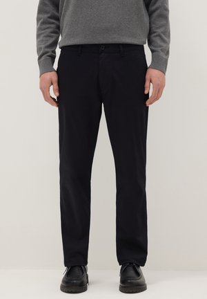 Homme portant un pantalon noir à jambes droites, des chaussures noires et une chemise grise à manches longues, debout devant un fond uni.