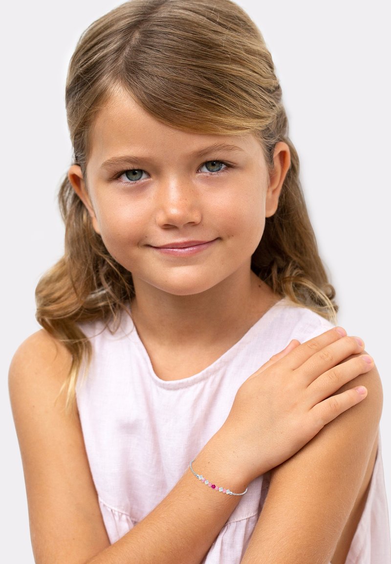 Pulsera de plata con piedras preciosas multicolores, que incluye piedras rosas, azules y transparentes, llevada en la muñeca de un niño. Top rosa claro.
