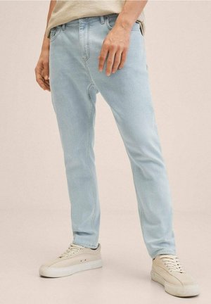 Jeans de mezclilla azul claro con un ajuste slim y un diseño clásico de 5 bolsillos, combinados con zapatillas blancas rotas sobre un fondo neutro.