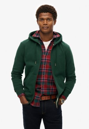 Green zip-up hoodie with a kangaroo pocket over a red and blue plaid shirt. Dark jeans and a brown belt complete the look.