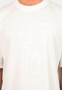 Weißes Baumwoll-T-Shirt mit Rundhalsausschnitt, kurzen Ärmeln und einem kleinen "DROPSIZE"-Logo in heller Farbe auf der linken Brustseite.