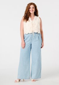Petit haut en crochet beige clair avec un devant noué et un ourlet festonné ; associé à un jean large en denim bleu moyen et des chaussures de couleur claire.