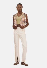 Chaleco bordado con patrones multicolores sobre una blusa verde clara, combinado con pantalones blancos con aberturas en los laterales y botones decorativos.