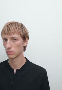 Sort ribbet knap-up skjorte med rund halsudskæring, båret af en model med kort, lysbrunt hår og lys hud, mod en lys baggrund.