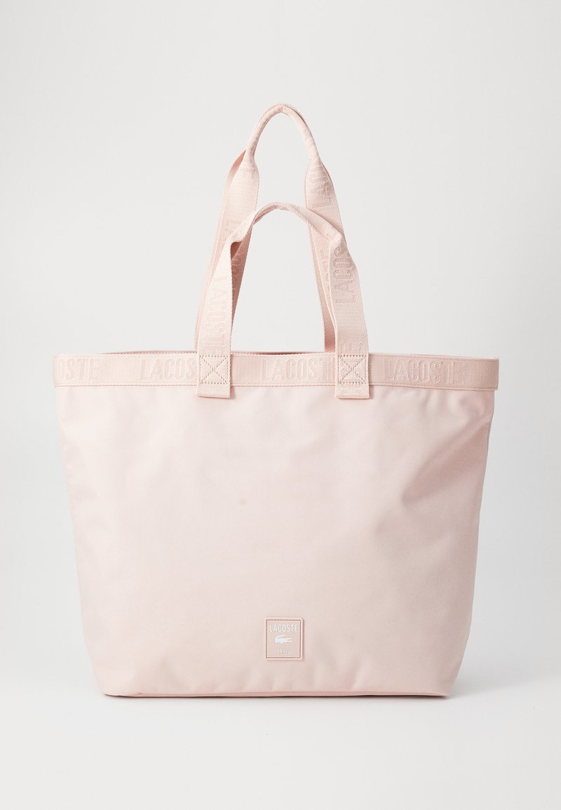 Borsa a tracolla Lacoste rosa chiaro con manici doppi e patch logo sul fronte, su uno sfondo bianco semplice.