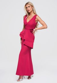 Femme blonde en robe rose fuchsia sans manches avec volants superposés et détail floral, portant des talons argentés et des boucles d'oreilles créoles, posant devant un fond blanc.
