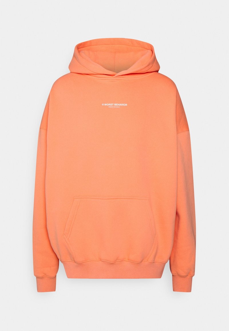 Sweat à capuche orange avec poche kangourou à l'avant et petit texte blanc "WORST BEHAVIOR" sur la poitrine.