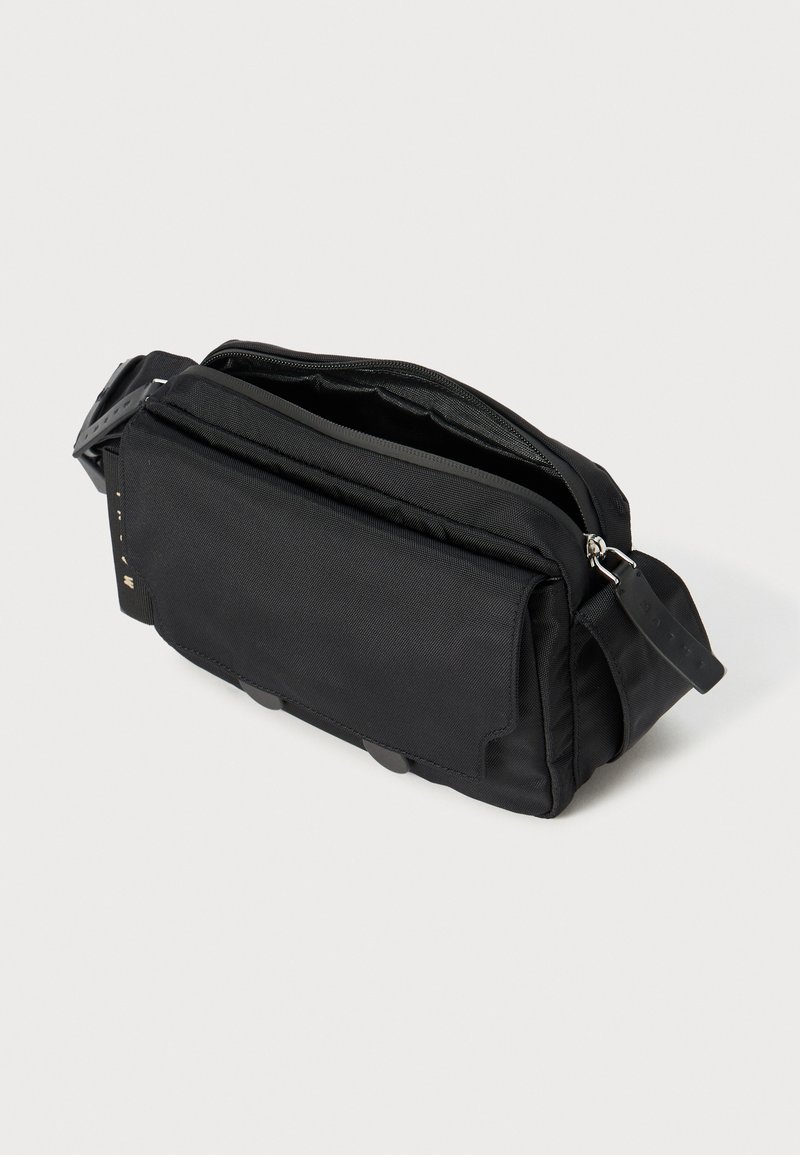Sac à bandoulière en tissu noir avec un compartiment principal zippé, une poche avant et une sangle ajustable ; doté d'une texture lisse et d'accents en métal circulaire.