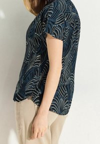 Camicetta navy con stampa astratta di foglie in crema. Maniche corte, stoffa morbida, vestibilità rilassata, orlo curvo e spacchi laterali. Abbinata a pantaloni beige.