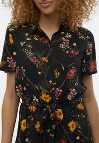 Kvinna med lockigt hår klädd i en svart blommig knapp-up klänning med knytbart midjebälte, prydd med gula, röda och rosa blommor.