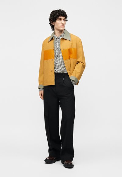 Άνδρας που φοράει μουσταρδί κίτρινο μπουφάν με πράσινο γιακά πάνω από ένα ασπρόμαυρο καρό πουκάμισο, μαύρο παντελόνι φαρδιάς γραμμής και σκούρα παπούτσια.