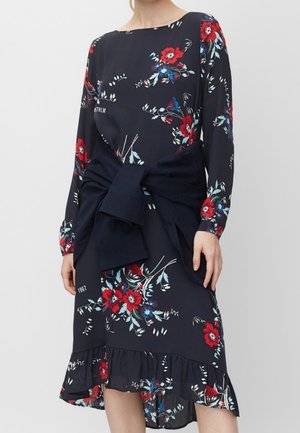 Robe fleurie marine avec des fleurs rouges et bleues, manches longues, ourlet à volants, et un pull marine noué autour de la taille.