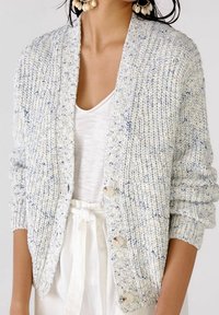 Kvinna iförd en prickig ljusblå grovstickad cardigan med stora knappar över en vit V-ringad topp och höga vita byxor med knyt.