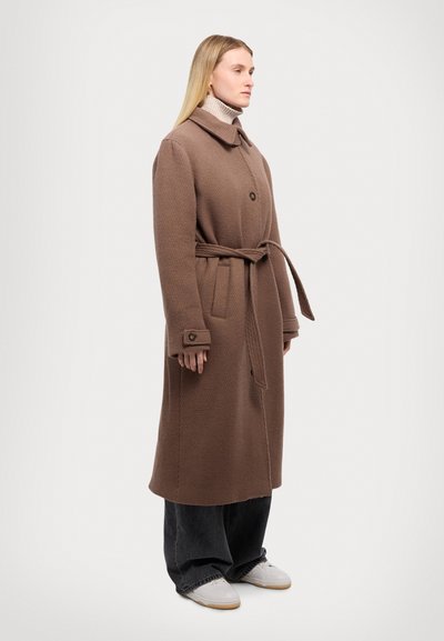 Brauner Fischgrätenmantel mit Gürtel, großem Kragen und Knopfleiste vorne. Getragen über einem hellen Rollkragenpullover und dunklen weiten Hosen. Weiße Sneakers.