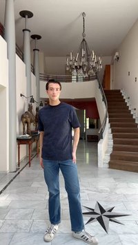 Ένας νεαρός άνδρας στέκεται ανέμελα σε ένα ευρύχωρο, φωτεινό δωμάτιο με μαρμάρινα δάπεδα, ένα μεγάλο πολυέλαιο και μια σκάλα με καφέ χαλί.