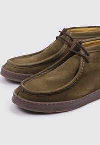 Zapatos de gamuza marrón con puntera redondeada, acentos cosidos y una suela de goma flexible. Presentan cordones marrones y un forro interior suave.