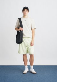 Tricou bej, geantă de umăr neagră, pantaloni scurți verzi deschis, adidași albi cu tălpi gri și șosete albe. Design simplu cu accente minime.