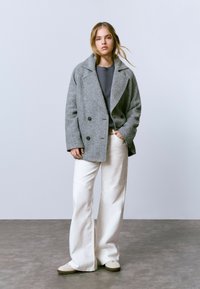 Una joven con el cabello rubio suelto lleva un abrigo gris de espiga de doble botonadura, una camisa oscura, unos pantalones anchos blancos y zapatillas beige, mientras está de pie sobre un suelo gris.