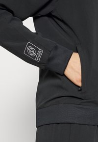 Person trägt schwarze Jacke mit hochgekrempeltem Ärmel, Hand in seitlicher Reißverschlusstasche und weiß gedrucktem Logo auf dem Unterarm.