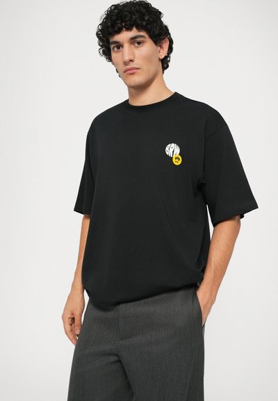 T-shirt nera a maniche corte in cotone, con una grafica bianca e gialla sul lato sinistro del petto. Il modello indossa pantaloni grigi.
